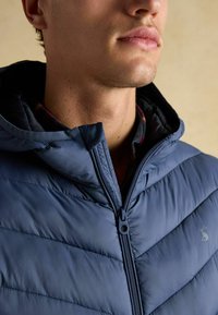 Mørkeblå quilted hættejakke med fuld lynlås og lysegrå logo på brystet. Indvendig foret med et ternet mønster.