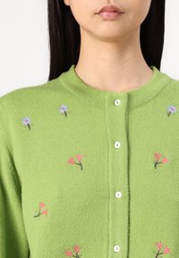 Grønn strikket cardigan med blomsterbroderi i rosa og blått, rund hals, og hvite knappelukkinger foran.