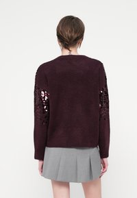 Burgunderfärgad tröja med långa ärmar, texturerad tyg och paljettdetaljer på axlarna. Kombinerad med en grå veckad kjol.