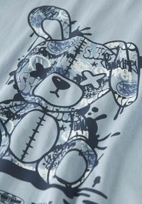T-shirt azzurro chiaro con grafica di un orso in blu scuro, decorato con un motivo cucito e accenti in stile graffiti. Realizzato in morbido cotone.
