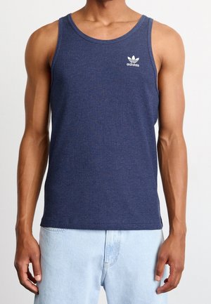 Mężczyzna ubrany w granatową koszulkę bez rękawów Adidas i jasnoniebieskie dżinsy, stojący na tle jednolitego białego tła.