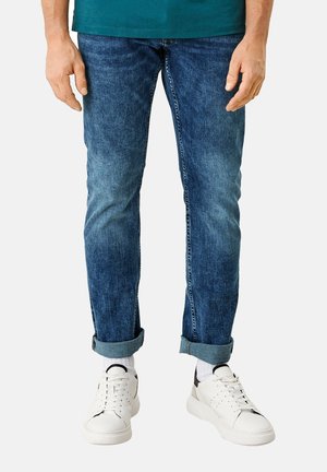 Homme portant un jean bleu retroussé, des chaussettes blanches et des baskets blanches, debout, les mains détendues le long du corps, devant un fond uni.