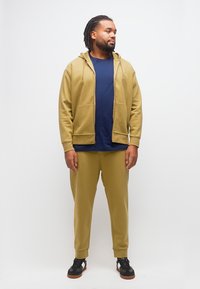 Ενήλικος άνδρας που στέκεται, φοράει φούτερ με φερμουάρ και κουκούλα σε ελιά πράσινο, ταιριαστά παντελόνια, μπλε σκούρα μπλούζα και μαύρα αθλητικά παπούτσια με λευκές λεπτομέρειες.