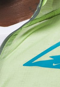 Lysegrøn vindjakke med tekstureret finish, med en fremtrædende blå Nike-logo foran og grå lynlåsdetaljer.