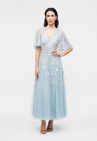 Kvinde står iført en lyseblå ankellang kjole med pink og hvid blomsterbroderi og korte flagrende ærmer, poserende med hånd på hoften.