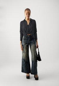 Crna skraćena bluza, plave široke traperice s patcima, i crne platforme; model drži malu crnu torbicu.