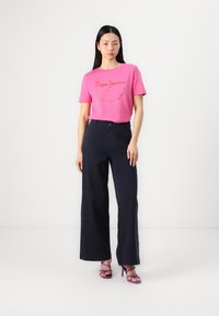 T-shirt de algodão rosa com logótipo em script, combinado com calças largas azul-marinho. O modelo usa sandálias roxas de salto. Design simples e moderno.