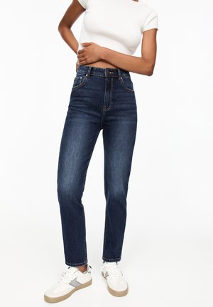 Woman wearing high-waist dark blue jeans, white short-sleeve crop top, and white sneakers with beige soles, standing against white background.