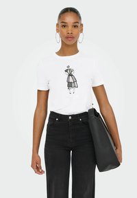 Hvid t-shirt med et sort grafisk design af en kvinde, parret med højtaljede sorte jeans. Modellen holder en sort tote bag.