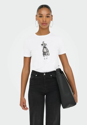 Weißes T-Shirt mit einem schwarzen grafischen Design einer Frau, kombiniert mit hochgeschnittenen schwarzen Jeans. Das Modell hält eine schwarze Tragetasche.