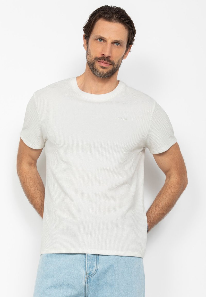 Uomo con barba che indossa una camicia bianca a maniche corte texture e jeans azzurri, in piedi con le mani dietro la schiena su sfondo uniforme.