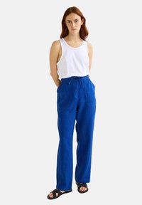 Pantalons en lin bleu avec une taille à cordon de serrage, deux poches avant et une coupe décontractée. Associés à un débardeur blanc uni et des sandales noires.