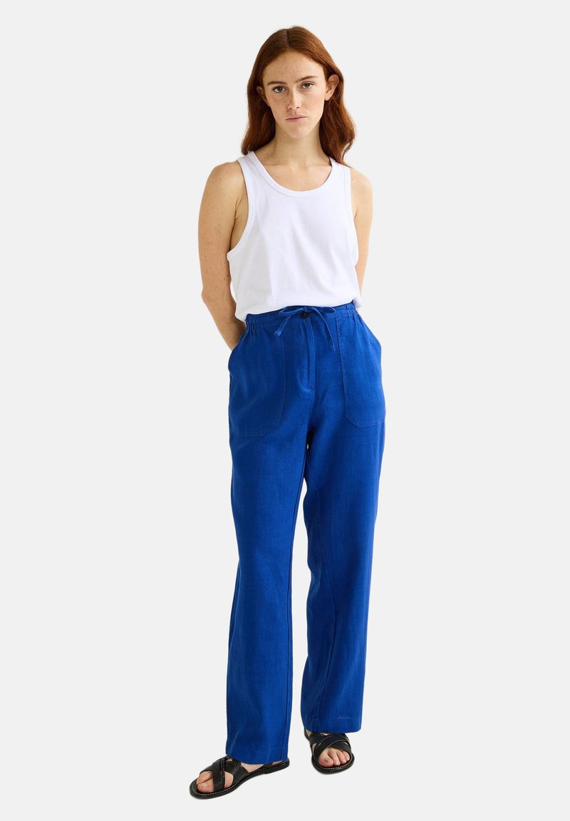 Pantalons en lin bleu avec une taille à cordon de serrage, deux poches avant et une coupe décontractée. Associés à un débardeur blanc uni et des sandales noires.