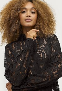 Zwarte kant blouse met bloemenpatronen, semi-transparant ontwerp, knoopdetails en een gestructureerde kraag. De textuur van de stof creëert een gelaagd effect.