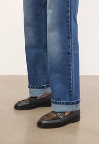 Mustad nahast loafid leopardimustriga detailiga, kantud siniste ülemise servaga teksadega sileda beeži põranda peal.