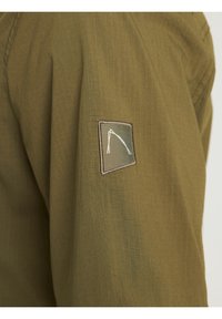 Oliwkowy materiał z prostokątną łatką, na której znajduje się stylizowana biała wędka na dwukolorowym zielono-brązowym tle.