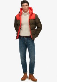 Daunenjacke mit orangefarbener Oberseite und olivgrüner Unterseite. Strickpullover in Cremefarbe darunter, blaue Jeans und braune Schnürstiefel.