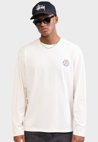 Uomo con berretto nero e occhiali da sole, camicia bianca a maniche lunghe con piccolo logo del globo e testo, e collana d'oro, su sfondo neutro.