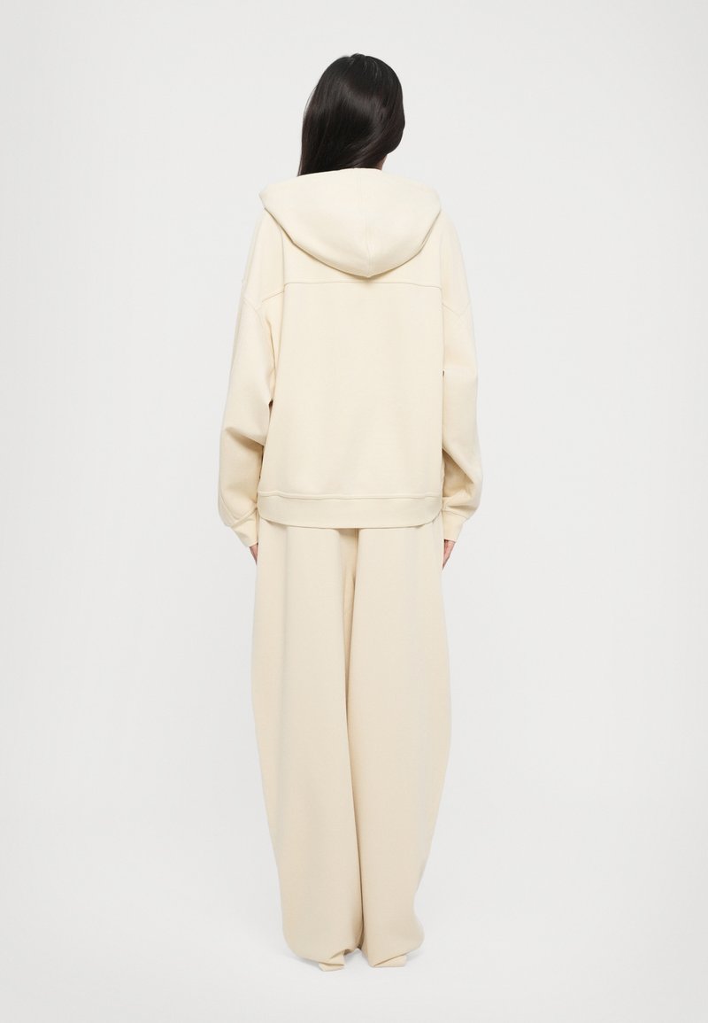 Felpa oversize color crema con cappuccio regolabile e maniche elastiche, abbinata a pantaloni a gamba larga sempre color crema, entrambi in tessuto morbido.