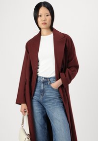 Abrigo burdeos de doble botonadura con solapas anchas, llevado sobre una blusa blanca ajustada y unos vaqueros azules de talle alto. Se sostiene un bolso de color claro.