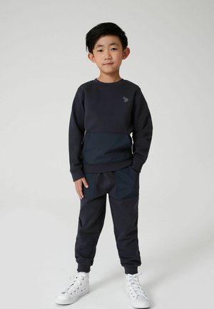 Felpa blu navy realizzata in tessuto testurizzato con dettagli scuri, abbinata a pantaloni della tuta coordinati con una tasca e polsini elasticizzati.