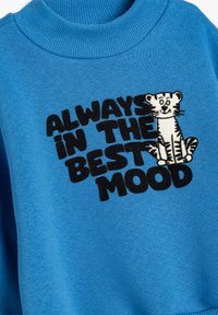 Blauer Sweatshirt mit geripptem Kragen, das schwarzen, gestickten Text "IMMER BESTE LAUNE" und eine Cartoon-Tiger-Illustration zeigt.