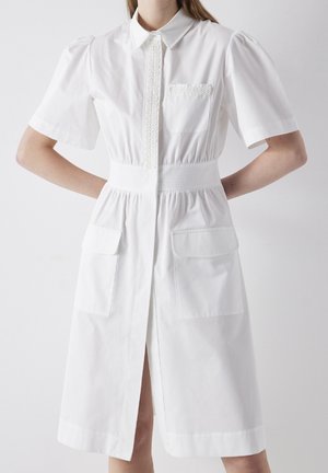 Valkoinen lyhythihainen mekko, jossa on liehuvat olkapäät, napituslistan pitsikoristelu, joustava vyötärö ja kaksi etutaskua. Kevyt puuvillakangas.