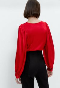 Blusa rossa di raso con maniche lunghe a sbuffo, scollatura tonda e dettagli arricciati. Abbinata a pantaloni neri a vita alta. Texture liscia.