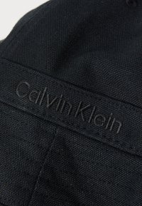 Crna platnena kapa s teksturiranim uzorkom i vezenom Calvin Klein logom na prednjoj strani. Ima zaobljeni vrh i ravni vizir.