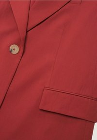Blazer rouge en tissu lisse, doté d'un revers cranté, d'une fermeture par un seul bouton et d'une poche passepoilée sur le côté gauche.