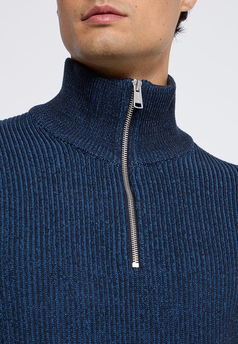 Dunkelblau gestrickter Pullover mit feinen vertikalen Streifen, hohem Kragen und silbernem Reißverschluss. Die gerippte Textur verleiht Tiefe.