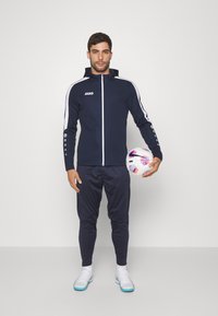 Marineblauwe trainingspak met witte strepen, rits aan de voorkant en capuchon. Model houdt een voetbal vast. Bevat een nauwsluitende broek en sportieve schoenen met lichte accenten.