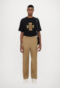 Maglietta grafica nera con design incrociato giallo e rosso, abbinata a pantaloni beige su misura e sneaker bianche. Sfondo semplice in monocromia.