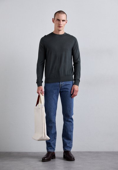 Dunkelgrauer langärmeliger Pullover, blaue Jeans, braune Lederstiefel, hält eine cremefarbene Tragetasche mit einem hellbraunen Griff. Schlichtes Design.