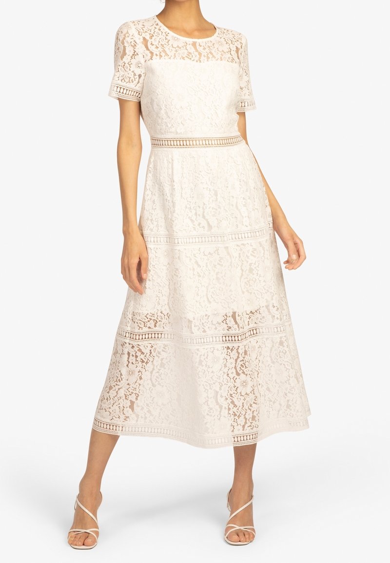 Femme portant une robe midi en dentelle blanche avec manches courtes et panneaux en dentelle transparente, associée à des talons blancs à brides.