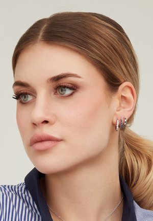 Femme aux cheveux châtain clair attachés en queue de cheval basse, portant trois boucles d'oreilles en argent en forme d'anneaux avec des pierres bleues et transparentes, une chemise rayée et un collier à chaîne en argent.