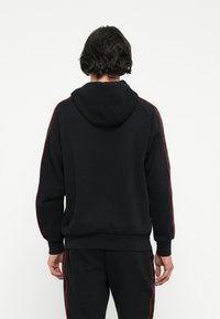 Sudadera con capucha negra con franjas rojas en los laterales, tejido texturizado y puños acanalados. Presenta un corte relajado y bolsillo tipo canguro.
