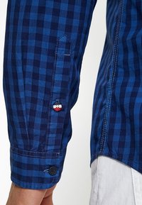 Niebieska koszula w kratę z różnymi odcieniami, mankiet zapinany na guzik z ozdobnym guzikiem oraz detal z naszywką na rękawie.