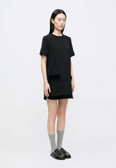 Joven mujer con un vestido corto negro con bolsillos, calcetas grises y mocasines negros, de pie frente a un fondo blanco sencillo.