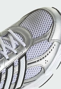 Ľahká atletická obuv so strieborným syntetickým prekrytím, bielou sieťovinou, čiernymi akcentovými pruhmi, bielymi šnúrkami a textúrovanou podrážkou.