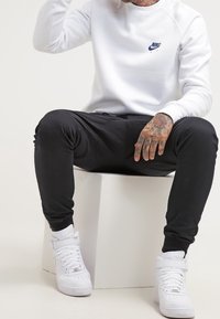 Witte Nike trui met blauw logo, zwarte joggingbroek en witte hoge sneakers. Model zittend op een witte kubus, met getatoeëerde handen zichtbaar.