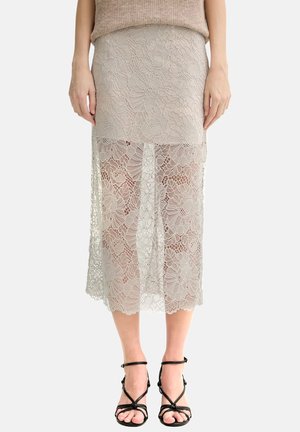 Mujer con falda midi de encaje beige sobre una combinación beige, combinada con sandalias de tacón negras con tiras, de pie frente a un fondo liso.