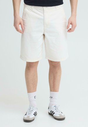 Mann trägt cremefarbene knielange Shorts, weiße Sportsocken und weiße Sneakers mit schwarzen Streifen und steht vor einem schlichten Hintergrund.
