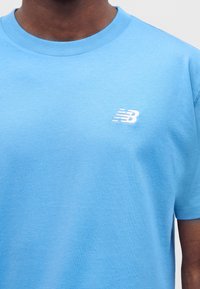 Άτομο που φοράει ανοιχτά μπλε μπλουζάκι New Balance με μικρό λευκό λογότυπο στο αριστερό στήθος, εμφανιζόμενο από τον λαιμό έως το μέσο του κορμού.