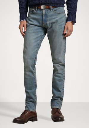 Homme portant un jean bleu clair, une ceinture en cuir marron, un pull bleu foncé et des chaussures en cuir marron à lacets, debout devant un fond uni.
