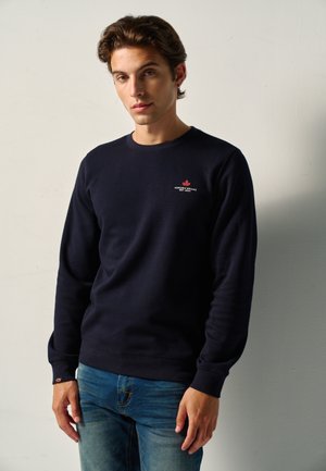 Joven con cabello corto y castaño que lleva una sudadera azul marino con un pequeño logo de hoja de arce roja y vaqueros azules frente a una pared lisa y clara.