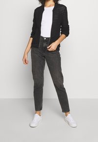 Cardigan en maille à rayures noires avec une fermeture éclair à l'avant, porté sur un t-shirt blanc, associé à un jean taille haute foncé et des baskets blanches.