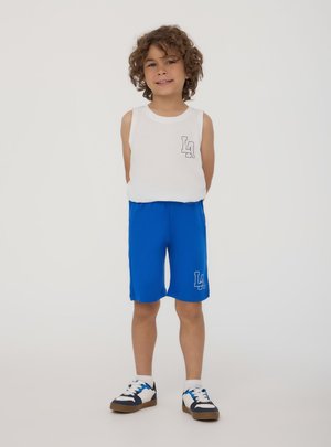 Giovane ragazzo con capelli ricci che indossa una canotta bianca e pantaloncini blu con loghi "LA", in piedi e sorridente su uno sfondo semplice.