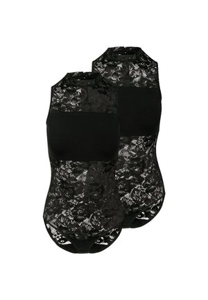 Femme aux cheveux bouclés portant un haut en dentelle noire sans manches et un short noir ample avec taille à cordon, debout devant un fond uni.