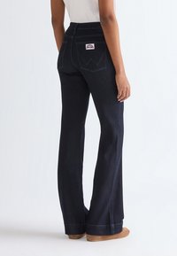 Person wearing high-waist dark blue wide-leg jeans with white stitching and a logo patch on the back pocket, standing barefoot.